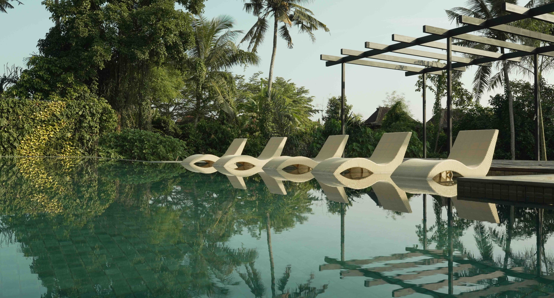Infinity pool with woven loungers reflected in calm water, surrounded by tropical palms at Kula Mani in Ubud, Bali